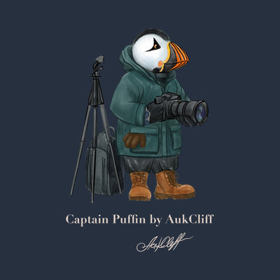 Captain Puffin in a green parka and walking boots, holding a camera beside a tripod