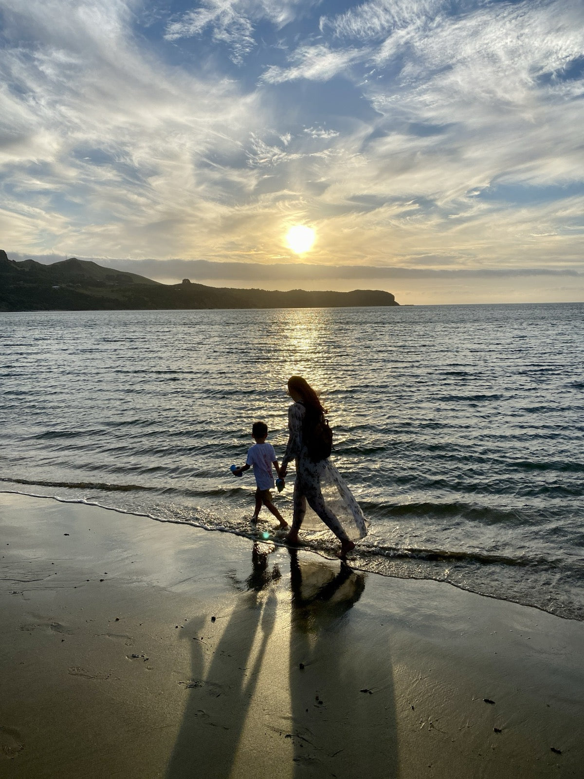 A parent and child walking the shoreline at sunset, hills across the bay